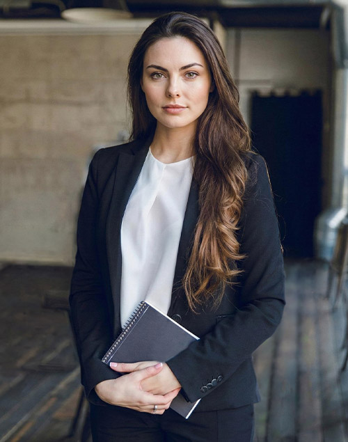 Eine Frau mit Bluse uns Blazer schaut in die Kamera und hält ein Notizbuch in der Hand.